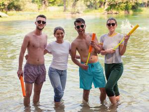 Eine Gruppe von vier jungen Menschen in sommerlichen Outfits steht fröhlich in einem Fluss.