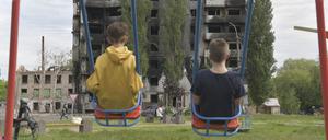 Zwei Kinder sitzen auf einer Schaukel und blicken auf ein durch russische Militärangriffe ausgebranntes mehrstöckiges Wohngebäude.