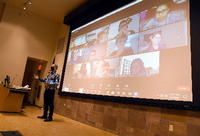 Ein Professor steht in einem Hörsaal, hinter ihm sind auf einer Leinwand Studierende zu sehen, die seine Vorlesung online verfolgen.