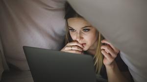Eine junge Frau schaut unter der Bettdecke liegend auf den Bildschirm ihres Laptops.