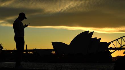 Ein Mann steht bei Sonnenuntergang vor dem Sydney Opera House und blickt auf sein Mobiltelefon. (Archivbild).