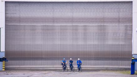 Werftarbeiter der Thyssen Krupp Marine Systems (TKMS) sind mit ihren Fahrrädern vor einem geschlossenen Tor einer Schiffbauhalle auf dem Gelände der Wismarer Werft unterwegs.