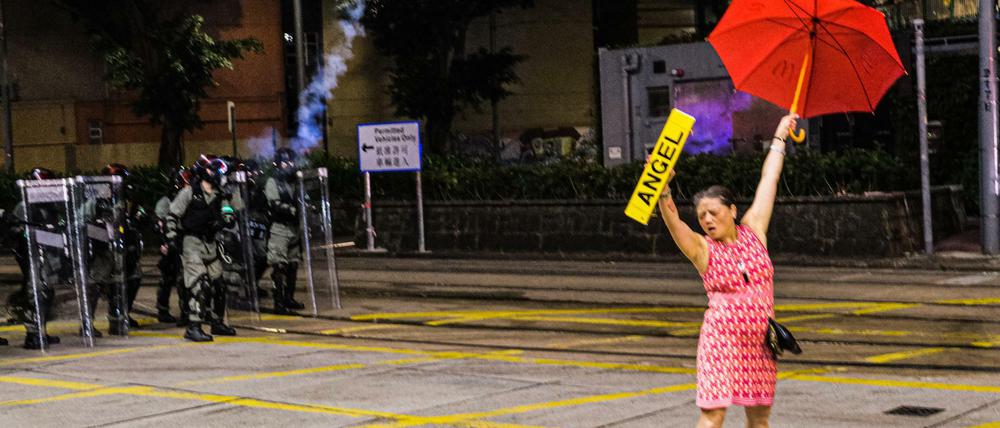 Eine Demonstrantin streckt einen Regenschirm und ein Schild in die Höhe, während Polizisten mit Tränengas auf sie zielen.