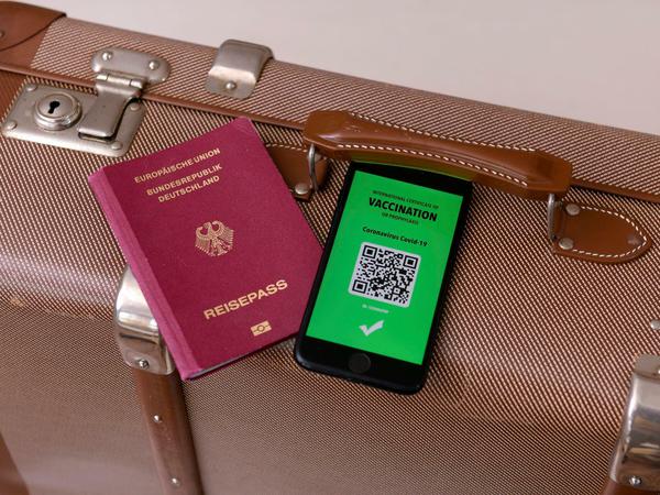 Ein imaginärer Impfpass in digitaler Form auf dem Smartphone iPhone, bereitgelegt auf einem Reisekoffer mit einem Reisepass.