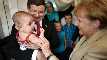 Bundeskanzlerin Angela Merkel und der türkische Ministerpräsident Ahmet Davutoglu unterhalten sich im Flüchtlingslager Nizip I mit einer Flüchtlingsfamilie, wobei Davutoglu das jüngste Kind auf dem Arm hält.