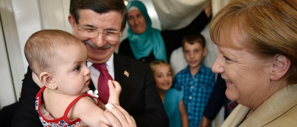 Bundeskanzlerin Angela Merkel und der türkische Ministerpräsident Ahmet Davutoglu unterhalten sich im Flüchtlingslager Nizip I mit einer Flüchtlingsfamilie, wobei Davutoglu das jüngste Kind auf dem Arm hält.