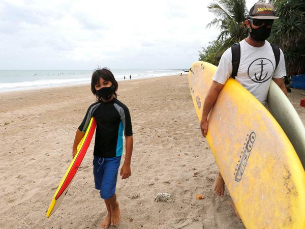 Indonesien, Bali: Touristen mit Mundschutz tragen ihre Surfbretter am Strand von Kuta.