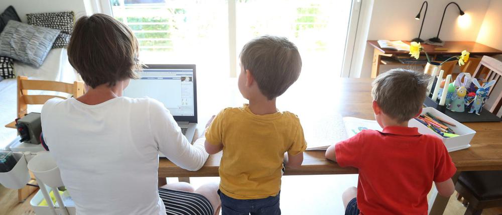 Die Mutter des sechsjährigen Jakob und des vierjährigen Valentin arbeitet Zuhause an einem Laptop, während ihre Kinder neben ihr malen und ein Buch ansehen. 
