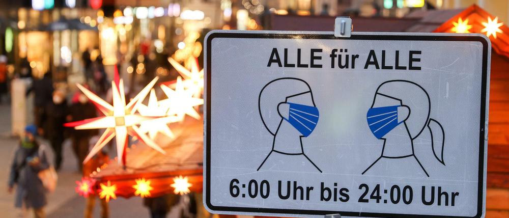 Passanten in Leipzig gehen hinter einem Schild entlang, das auf die Maskenpflicht in der Fußgängerzone hinweist.