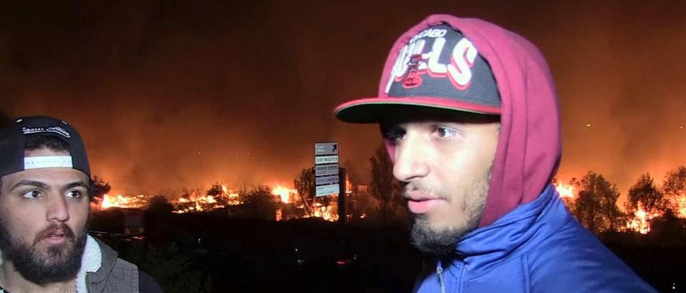 EinFlüchtling aus dem Irak (r) spricht mit Journalisten, während im Hintergrund ein Flüchtlingslager in Flammen steht.