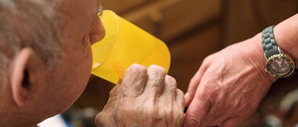 Eine ambulante Pflegerin hilft einem alten Mann bei der Tabletteneinnahme, der Mann trinkt Wasser. 