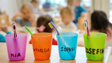 Zahnbürsten und Zahnputzbecher mit den Vornamen der Kinder stehen in einer Kindertagesstätte auf einem Tisch. 