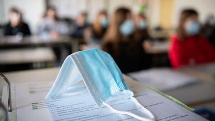 Eine Maske liegt im Unterricht auf Unterlagen, während im Hintergrund Schüler:innen mit Mund- und Nasenschutz zu sehen sind.