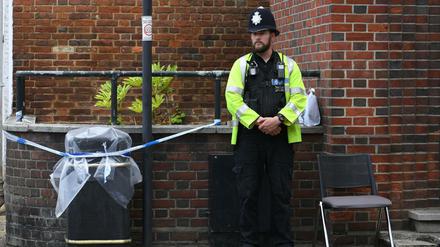 Ein Polizist steht vor einem mit Polizeiband abgesperrten und Plastikfolie bedeckten Mülleimer.
