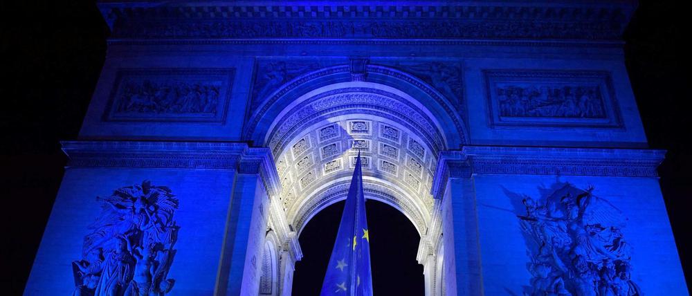 Menschen versammeln sich vor dem blau beleuchteten Arc de Triomphe in Paris, in dessen Bogen eine EU-Flagge hängt