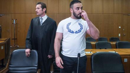 Mit einem weißen T-Shirt bekleidet steht der Angeklagte Aria L. in einen Hochsicherheitssaal des Oberlandesgerichts in Frankfurt am Main neben seinem Anwalt Andreas Bensch.
