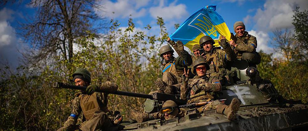 Ukrainische Soldaten gestikulieren und halten eine ukrainische Fahne, während sie auf einem gepanzerten Fahrzeug eine Straße zwischen Isjum und Lyman entlang fahren.