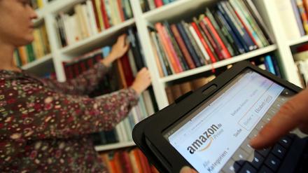 Auf einem Tablet-Computer sucht ein Mann bei dem Online-Versandhaus Amazon nach Literatur, während seine Frau ein Buch aus dem Bücherregal nimmt. 
