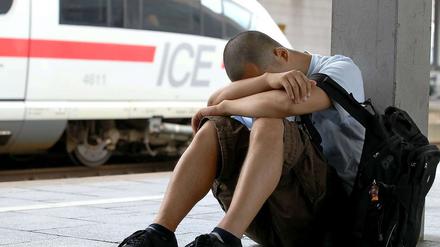 Ein junger Mann in kurzer Hose sitzt auf einem Bahnsteig, den Kopf auf die Arme gestützt. Im Hintergrund steht ein ICE-Zug.