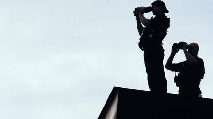 Zwei bewaffnete Männer mit riesigen Ferngläsern stehen im Gegenlicht auf einem Dach.