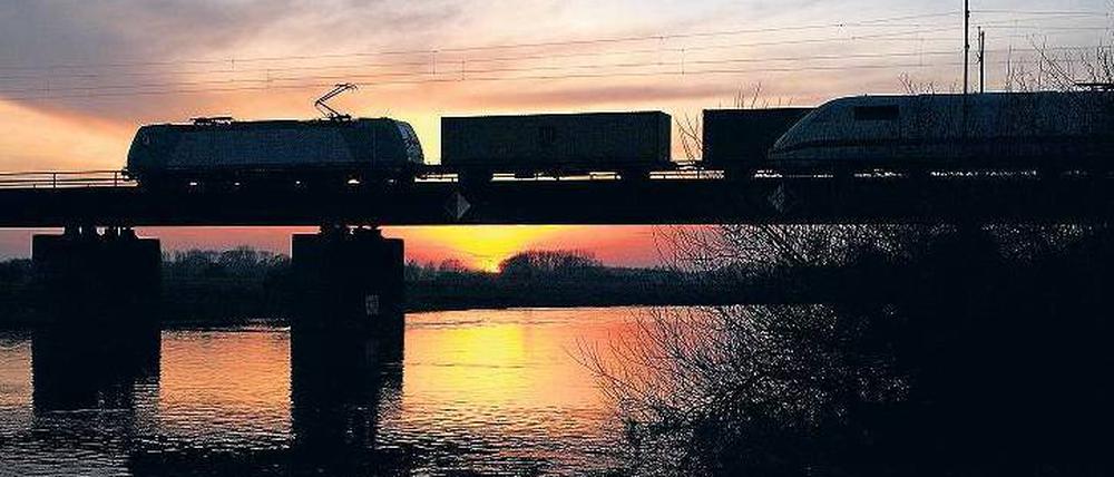 Ein Containerzug fährt bei Sonnenuntergang über eine Aller-Brücke und wird von einem ICE überholt.