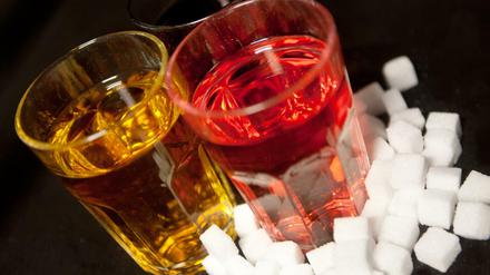 Drei Gläser gefüllt mit roter Limonade, Cola und einem Energy-Drink stehen neben Zuckerwürfeln auf einem Tisch. 