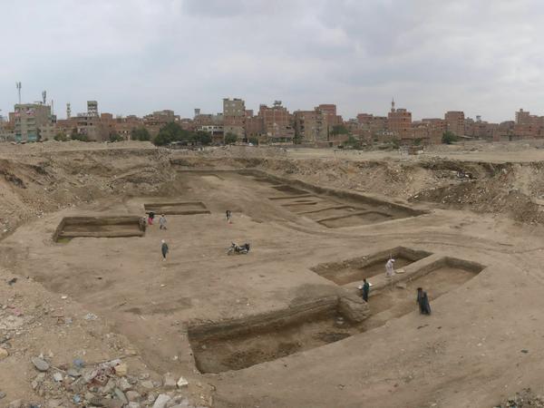 Eine archäologische Grabung mit mehreren Vertiefungen im Boden, im Hintergrund sind moderne Hochhäuser zu sehen.