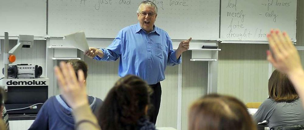 Ein Lehrer steht gestikulierend und lachend vor einer Schulklasse, die Schüler melden sich.