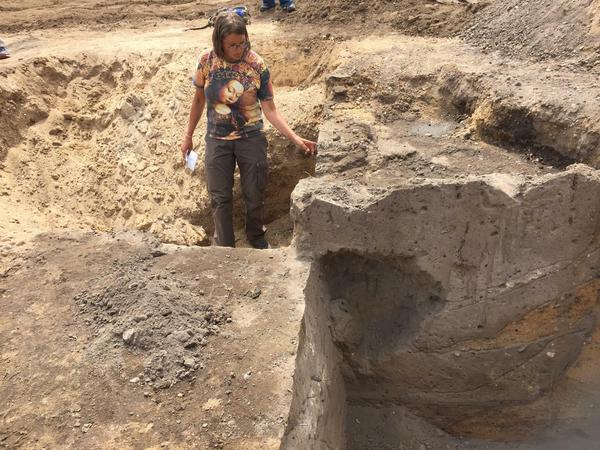 Eine Archäologin steht am Rand einer Grabung, in deren Querschnitt eine große Ausbuchtung zu sehen ist.