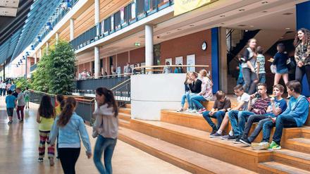 Das frisch sanierte Foyer einer Ganztagsschule, in dem Schüler auf Holzstufen sitzen und Schülerinnen an begrünten Flächen vorbeigehen.