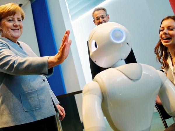 Beim Girls Day 2017 interagiert Angela Merkel mit einem Roboter, eine junge Frau schaut lachend zu.