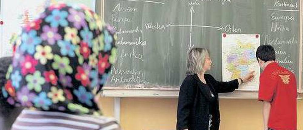 Eine Lehrerin erklärt einem Schüler an der Tafel eine Landkarte. Im Vordergrund sitzen ein weiterer Schüler mit dunkler Hautfarbe und eine Schülerin mit Kopftuch.