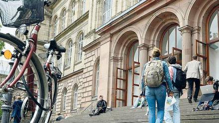 Studierende betreten ein Universitätsgebäude, einige sitzen auf der Treppe.