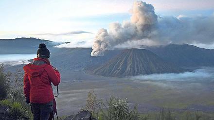 Eine Frau steht auf einer Anhöhe und schaut in Richtung eines aktiven Vulkans, über dem eine Rauchwolke steht.