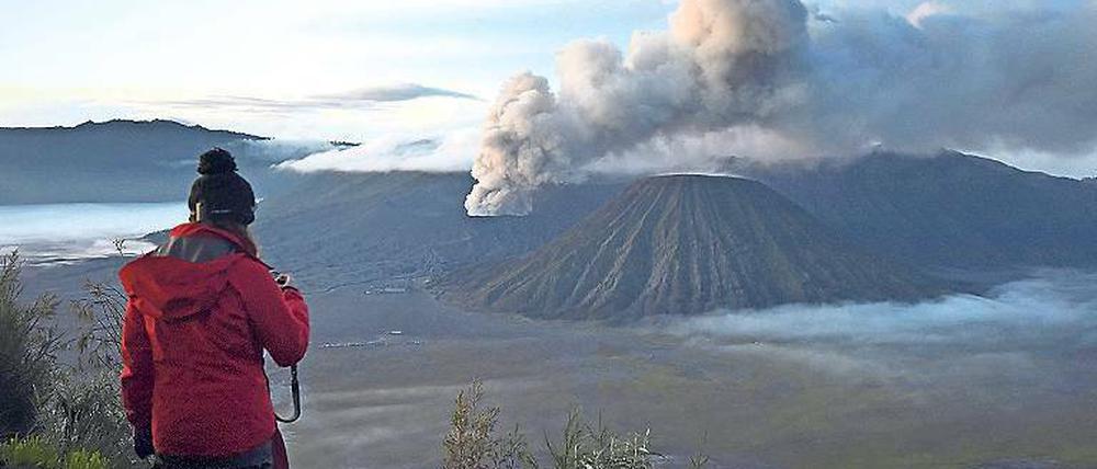 Eine Frau steht auf einer Anhöhe und schaut in Richtung eines aktiven Vulkans, über dem eine Rauchwolke steht.