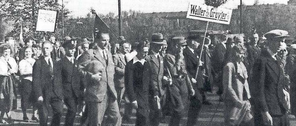 Männer und Frauen marschieren über einen Platz, dabei halten sie ein Schild mit der Aufschrift Walther de Gruyter hoch.