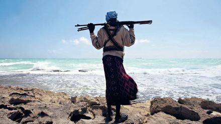 Ein Mann, der ein Gewehr über den Schultern trägt, steht auf einem felsigen Ufer und schaut über das Meer.