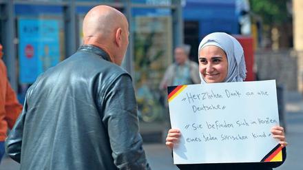 Eine Frau mit Kopftuch hält einem älteren Mann ein Schild entgegen, die Aufschrift besagt unter anderem: Herzlichen Dank an alle Deutsche.