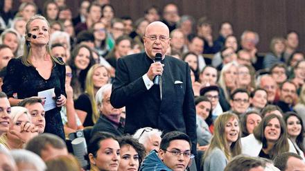 Ein Mann mit einem Mikrofon in der Hand steht inmitten von Studierenden, die in einem Hörsaal sitzen.