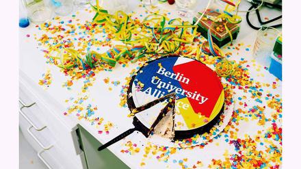 Eine Torte mit der Aufschrift Berlin University Alliance steht auf einem Schreibtisch, der mit Luftschlangen und Konfetti bedeckt ist.