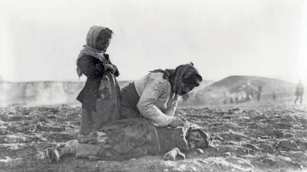 Auf einem unbestellten Feld beugt sich eine Frau über ein am Boden liegendes Kind, ein zweites Kind steht andächtig daneben.