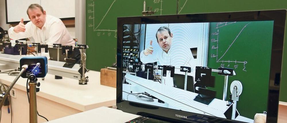 Ein Professor ist bei einer Physik-Vorlesung im Hörsaal live und auf einem Bildschirm zu sehen.