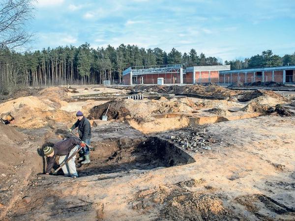 Zwei Archäologen graben zwischen einem Waldstück und dem Gebäude einer Gedenkstätte nach Spuren der ehemaligen Lagerinsassen.