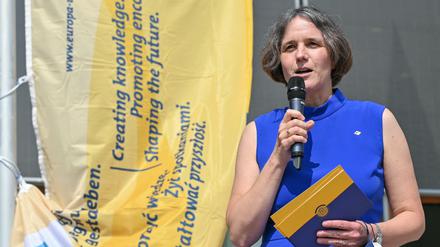 Von Blumenthal steht in einem blauen Kleid vor einem Banner mit blauer Schrift auf gelbem Grund und spricht in ein Mikrofon.