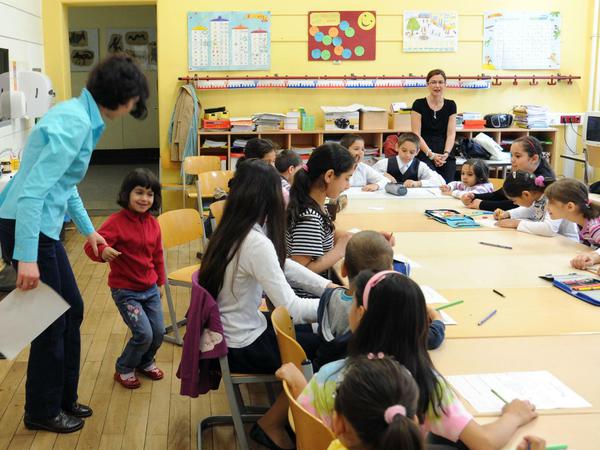 Jungen und Mädchen sitzen in einem Klassenraum an einem großen Tisch und machen unter der Aufsicht von Lehrerinnen Schreibübungen.