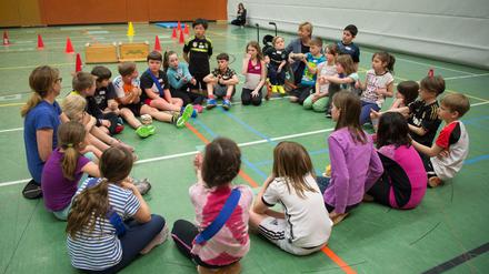 Lehrkräfte und Schulkinder in Sportbekleidung sitzen in einer Turnhalle in einem Kreis und sprechen miteinander. Ein Kind sitzt im Rollstuhl.