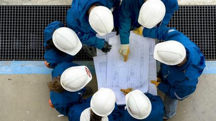 Eine Gruppe von Ingenieuren mit Bauhelmen auf dem Kopf beugt sich über Baupläne.