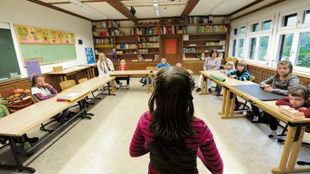 Eine Schülerin im Grundschulalter steht in einem Klassenraum vor neun Mitschülern verschiedenen Alters und zwei Lehrerinnen.