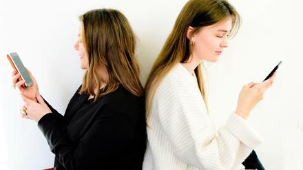 Zwei Frauen sitzen Rücken an Rücken und tippen Sprachnachrichten in ihre Handys ein.