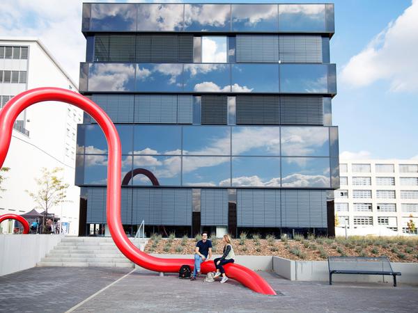 Auf dem HTW-Campus sitzen zwei Studierende auf einer Skulptur vor einem Gebäude mit spiegelnder Fassade.